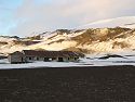 An old building on Deception Island is in shade as the long sunset lights the slopes above.